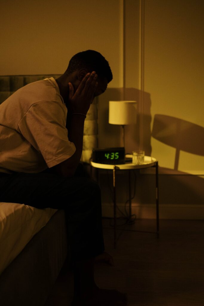 Man sitting on the edge of a bed with his head in his hands. Alarm clock showing 4:35 am. 