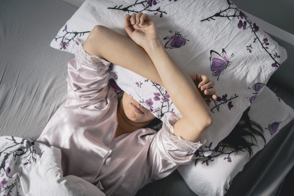 Lady lying in bed with her hands behind her head and a pillow over her eyes 